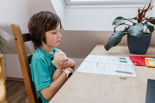 Child doing homework at desk hugging toy indoors