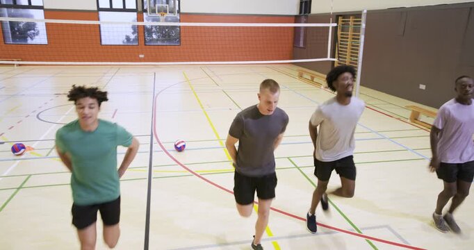 Five men starting warmup, jogging with high knees at school gym under net, avoiding volleyball