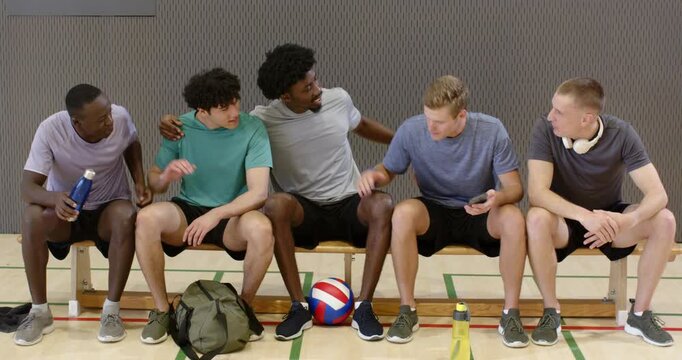 Five male teammates sitting on bench in gym, placing volleyball on floor, linking arms during break