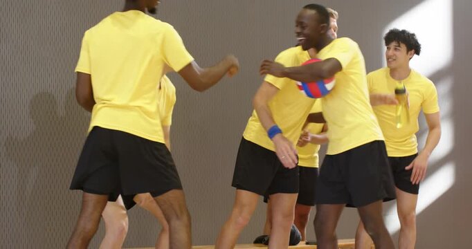 Diverse male volleyball team in yellow approaching bench greeting, passing ball at gym cheering