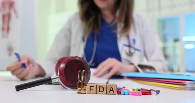 Wooden blocks spell abbreviation FDA near gavel and capsule tablets. Doctor woman reviews safety file signing approval note for new drug batch