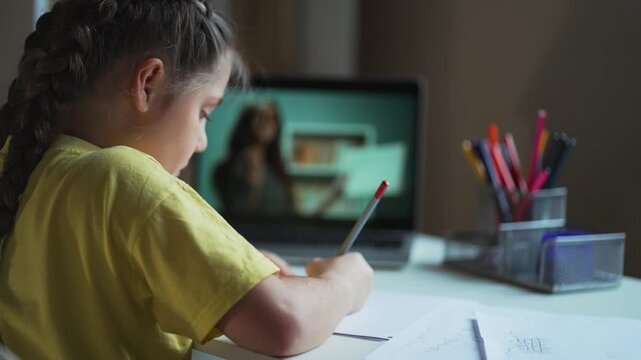 Child writing homework at desk focused on study with pencil and paper while student watches teacher on laptop during online class showing learning and concentration in homeschool environment quiet