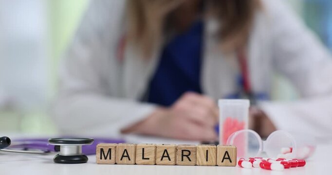 Wooden blocks spell word Malaria near capsules and stethoscope on desk. Doctor woman writes prescription reviewing patient chart for fever control
