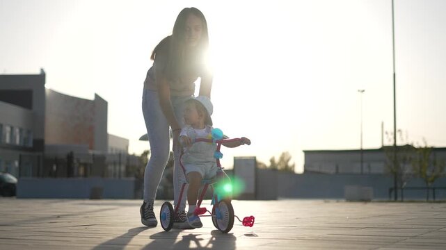 Helping toddler learn to ride mother guiding child on tricycle in urban park at sunset, parent teaching balance and confidence, family time, soft backlight, playful interaction with training wheel