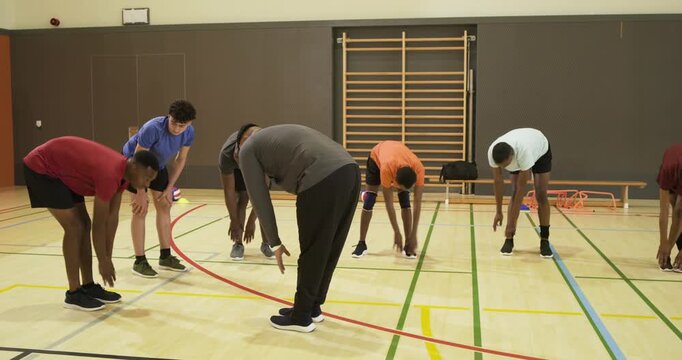 Seven male team warming up in school gym after coach showing stretch and correcting near volleyball