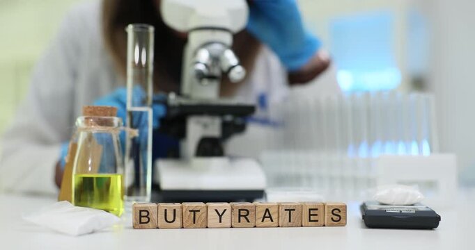 Wooden cubes neatly arranged to form word Butyrates identifying substance under investigation. Chemist examines substance through microscope by tubes
