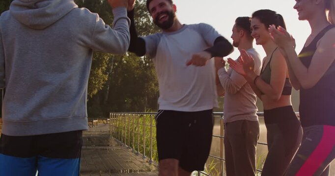 Happy athletes of fitness team celebrate goal achievement and triumph of marathon runner in summer park. People applaud running winner, give high five to man after race and success of run outdoor