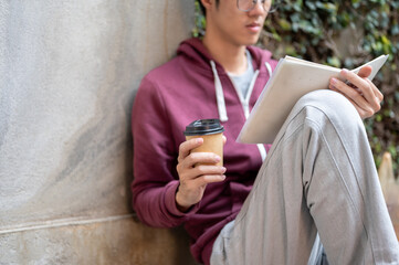 Fototapeta premium Glasses young asian man holding coffee reading a book while sitting knee up leaning on concrete wall. Public Space, Leisure Time, Chilling Outdoors.