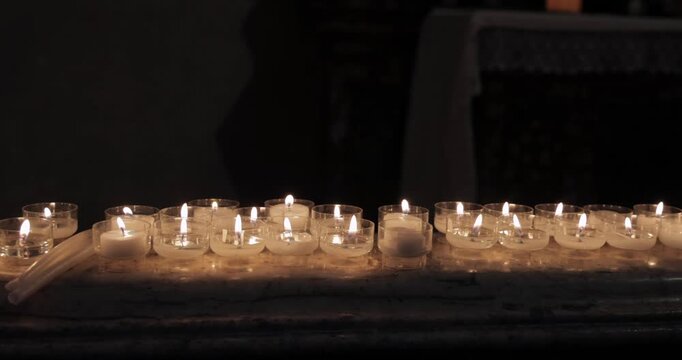 Candlelit prayer candles on marble ledge in a dim church, camera tracks along a row of tiny flames as wax pools in clear cups and warm reflections shimmer across stone while deep shadows