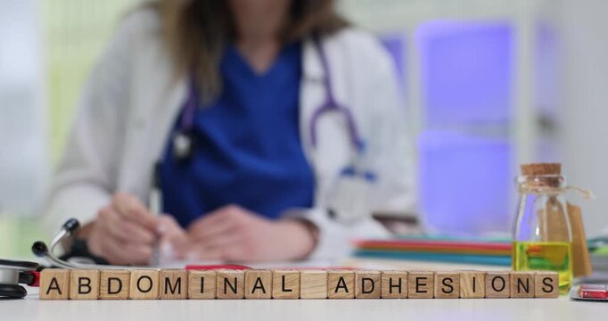 Miniature wooden cubes arranged on desk form phrase Abdominal Adhesions. Young woman doctor reviews notes and writes treatment plan for patient