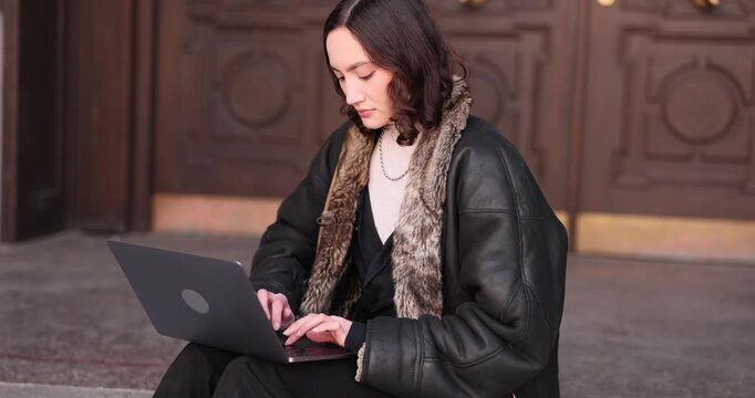 Woman freelancer sits comfortably on steps near door and works on modern laptop. Female types document as part of freelance work. Slow motion