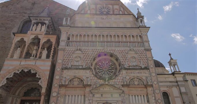Colleoni Chapel facade in Bergamo Italy, low angle tilt down reveals roof gable then rose window and carved portal as midday sun starburst and lens flare glow over marble patterns