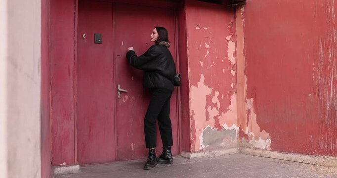 Woman in autumn clothing stands by metal door of old apartment entrance and knocks. Female tilts head slightly to catch response. Slow motion