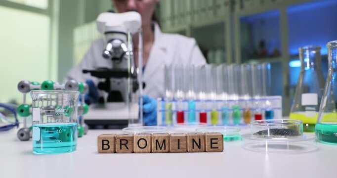 Wooden blocks spell word Bromine near microscope and colored liquids. Lab researcher handles glassware preparing halogen samples for experiment