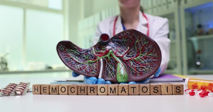 Wooden blocks spell Hemochromatosis near liver model and pill blisters. Doctor holds organ replica explaining iron overload risk during checkup