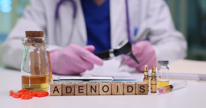 Wooden blocks spell Adenoids near vials and medical tools on desk. Doctor prepares examination instrument writing care plan for airway problem