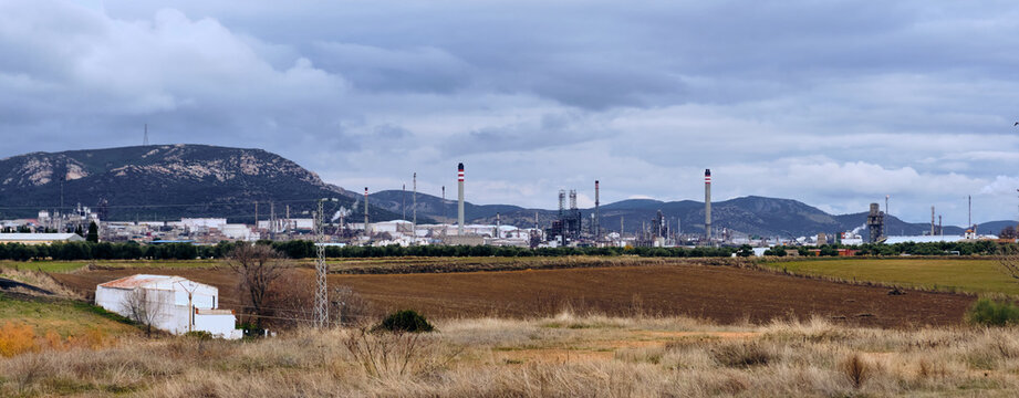 View of a sprawling industrial complex nestled against rolling hills under a brooding sky, contrasting with the earthy tones of the fields, Puertollano, Castilla-La Mancha, Spain.