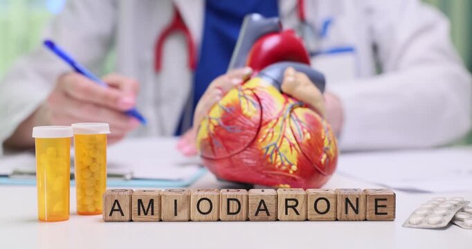 Wooden blocks spell word Amiodarone near heart model and pill bottles. Cardiologist reviews case preparing antiarrhythmic dose for patient safety