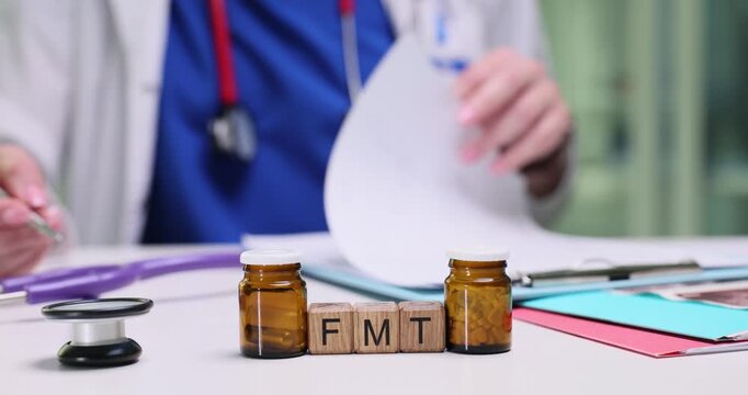 Wooden blocks spell abbreviation FMT between amber jars and stethoscope. Doctor labels specimen containers filing case notes for microbiome therapy