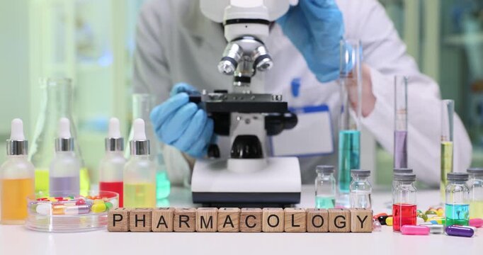 Wooden blocks spell word Pharmacology near microscope and droppers. Lab worker studies medicine samples and tablets recording results for therapy