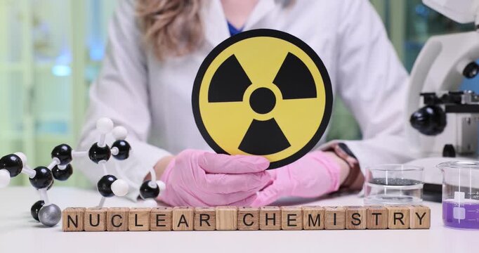 Wooden blocks spell phrase Nuclear Chemistry near radiation symbol and lab glassware. Scientist wears gloves reviewing compounds holding warning sign