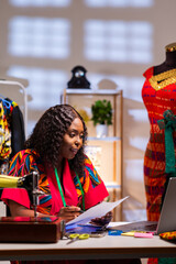 Fototapeta premium African fashion designer managing business, reviewing documents, and using laptop in her studio