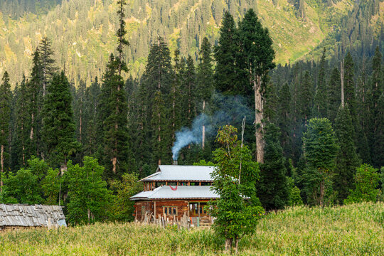 View of a rustic cabin nestled amidst the lush green meadow and towering trees, with smoke rising from the chimney in Arang Kel, Azad Kashmir, Pakistan.