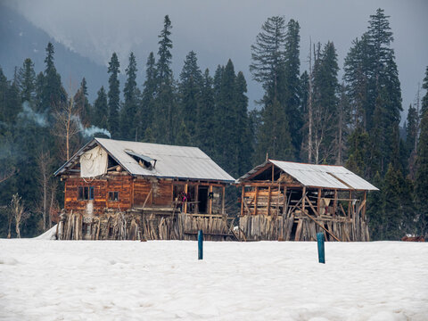 View of rustic wooden houses stand amidst a snow-covered landscape, smoke curling into the crisp air, framed by towering pine trees, Arang Kel, Pakistan.