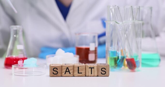 Wooden blocks spell word Salts on lab table near glassware. Scientist measures colored liquids preparing samples for chemical testing and analysis