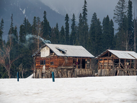View of rustic wooden huts nestled in a snow-covered landscape, smoke rising against a backdrop of towering evergreens, creating a serene winter scene, Arang Kel, Pakistan.