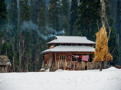 View of a rustic wooden house with smoke gently rising from its chimney, nestled amidst snow-covered ground and towering evergreen trees, Arang Kel, Pakistan.