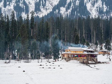 View of wooden buildings nestled in a snowy valley, smoke rising against a backdrop of dense evergreens and snow-capped mountains, Arang Kel, Pakistan.