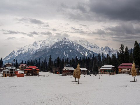 View of snow-covered village nestled against towering, snow-capped mountains under a cloudy sky, evergreen trees dotting the landscape, Arang Kel, Pakistan.