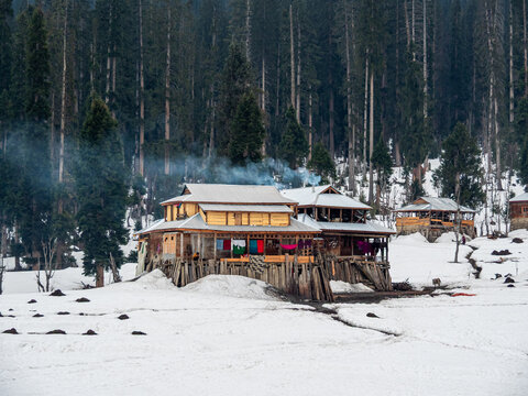View of wooden houses standing in stark contrast to the pristine white snow, smoke curling upwards against a backdrop of dense, dark evergreen forest, Arang Kel, Pakistan.