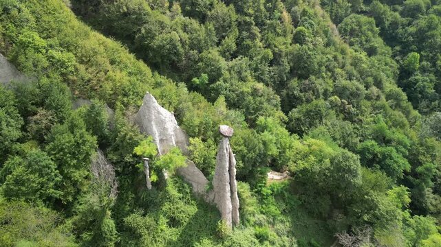 Zone earth pyramids capstone spire flyover (Piramidi di Zone) near Lake Iseo Italy, drone sweeps above forested gorge to spotlight a huge boulder perched on a tall clay hoodoo