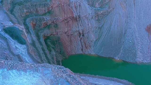 Descending quarry basin filling with green water beneath steep terraced rock walls. Curving open pit excavation collecting water along layered mineral slopes. Forming artificial lake inside deep
