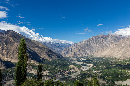 Aerial view of rugged, rocky mountains meeting a lush green valley, crowned with snow-capped peaks under a crisp blue sky, Hunza Nagar, Gilgit, Pakistan.
