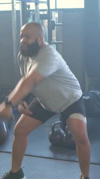 Vertical video: Reaching man grabbing kettlebell and swinging in gray tee for set at gym with rack