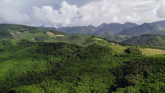 Sideward panning motion flying view of hills and valleys covered with lush dense tropical foliage, with hazy mountainous landscape and cloudy sky in the background.
