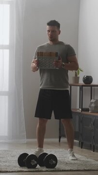 Man exercising at home holding heavy basket of books while standing near dumbbells in bright living room, demonstrating improvised strength workout routine
