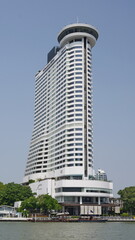 Fototapeta premium The Millennium Hilton hotel featuring its distinctive circular rooftop 360 bar stands along the Chao Phraya river under a clear sky in Thai capital Bangkok