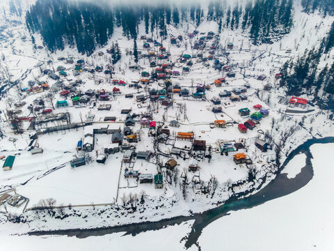 Aerial view of snow-blanketed rooftops punctuate the winter landscape, nestled beside the winding river beneath the misty mountain peaks, Sharda, Azad Kashmir, Pakistan.