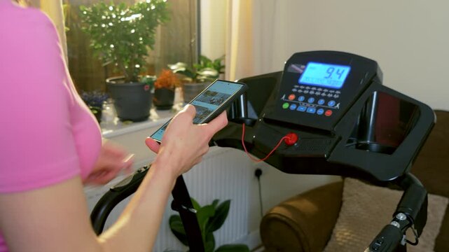 Scrolling smartphone gallery while running on treadmill unrecognizable woman during indoor home cardio session. Using mobile phone while jogging on electric running machine near window plants in