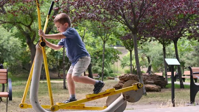 A preschool boy uses an outdoor elliptical trainer in a park during the summer.