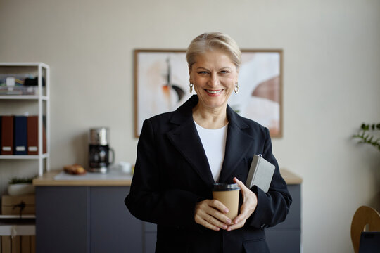 Portrait of senior businesswoman holding takeaway coffee and notebook, smiling at camera in office. Ideal for leadership, executive branding, mentoring, career success marketing