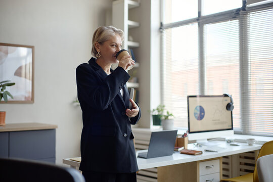 Senior businesswoman drinking takeaway coffee in office, pausing during workday near computer setup. Suitable for corporate productivity, leadership, office routine marketing