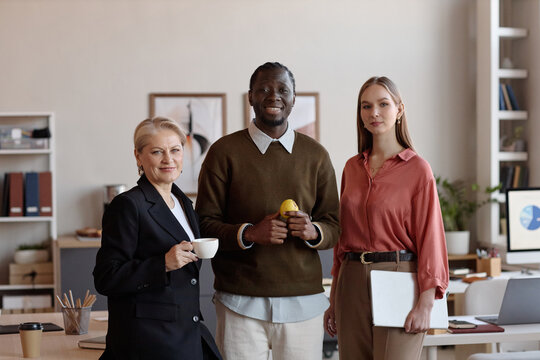 Portrait of diverse business team standing in office, smiling at camera and holding coffee, fruit, laptop. Suitable for corporate communication, teamwork, leadership, hiring campaigns