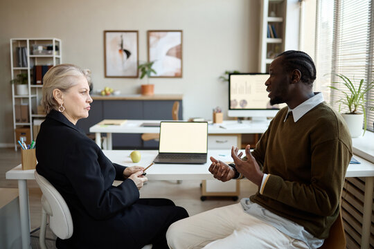 Black young adult man discussing work strategy with senior businesswoman in office meeting. Useful for corporate communication, mentoring, leadership, teamwork marketing
