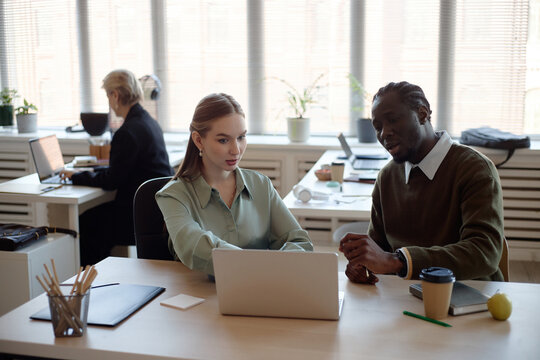 Young Caucasian businesswoman and Black businessman reviewing laptop data, discussing project strategy in office. Ideal for teamwork, corporate training, collaboration, medium shot