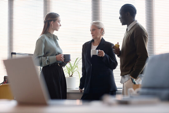 Young adult Caucasian woman, senior woman, Black man discussing work during office coffee break, holding mugs and snack. Suitable for teamwork, corporate culture, staff communication